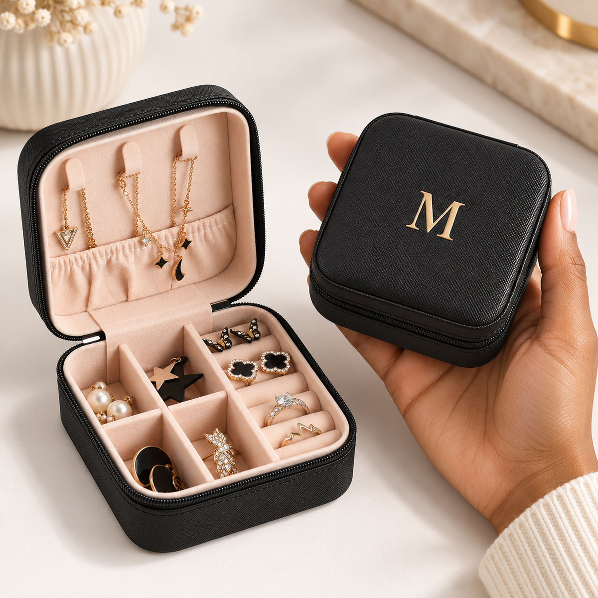 A black zippered jewelry organizer case is shown open with pink interior compartments holding earrings and rings, alongside a hand holding the closed case which has a gold monogram letter 'M' on top. The background is a clean, light-colored tabletop with soft-focus decorative elements.