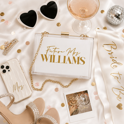 A styled flat-lay of a clear acrylic clutch bag with a gold chain strap, personalized with 'Future Mrs. WILLIAMS' in gold lettering on a white front.