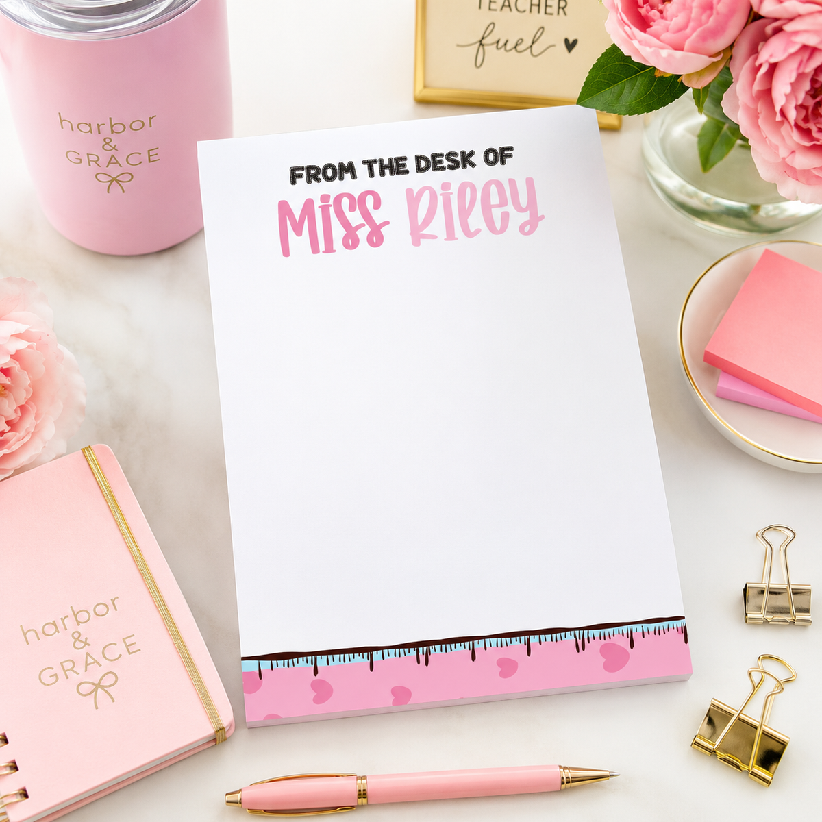 A white notepad with pink and black text reading 'FROM THE DESK OF Miss Richey' is styled on a light surface with pink stationery items, including a pink notebook, pen, clips, and flowers.